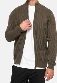 Olive green zip-up jacket with a textured zigzag pattern, ribbed cuffs and collar, worn over a white t-shirt, paired with black pants.