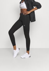 Svarta leggings med en åtsittande design, med en vit Puma-logotyp och reflektiva detaljer, tillsammans med vita sportskor.