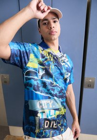 Korte mouw grafisch shirt met levendige blauwe en gele patronen, met tekstelementen en een polo kraag. Gestyled met een witte pet.