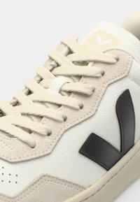 Sneaker featuring a white leather upper, beige suede accents, black logo, and beige laces. Textured design with perforations on the toe.
