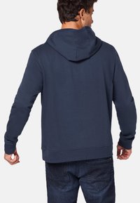 Mörkblå huvtröja med långa ärmar, ribbade muddlinningar och en mjuk textur. Har en kenguruficka och en rak fåll.