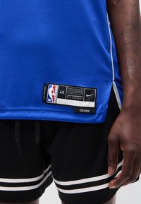 Κοντινό πλάνο ενός ατόμου που φοράει μπλε αυθεντική φανέλα NBA Authentic swingman και μαύρο σορτς με λευκές ρίγες, δείχνοντας την ετικέτα της φανέλας και το τατουάζ στο αντιβράχιο.