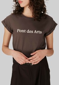 T-shirt en coton marron avec des manches roulées, présentant le texte blanc "Pont des Arts" sur le devant. Texture lisse, coupe décontractée.
