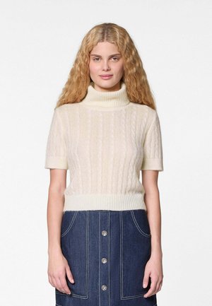 Frau mit langen lockigen blonden Haaren, die einen cremefarbenen, kurzärmeligen Rollkragenpullover mit Zopfmuster und einen dunkelblauen Jeansrock mit Knopfleiste trägt.