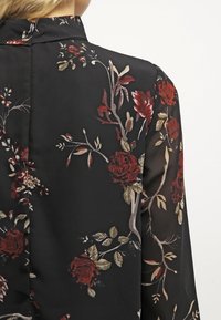 Blusa nera floreale con rose rosse e dorate e foglie verdi, realizzata in tessuto trasparente, con colletto alto e un design posteriore sobrio.