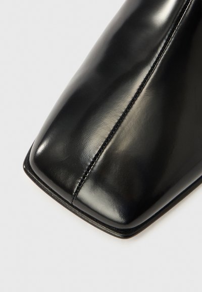 Μαύρο δερμάτινο παπούτσι με τετράγωνη μύτη, λείο φινίρισμα και λεπτομέρειες διακριτής ραφής κατά μήκος της πλευράς. Η σόλα είναι λεπτή και ελαφρώς ανυψωμένη.