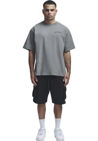 Jeune homme debout, portant un T-shirt gris oversize, un short cargo noir, des chaussettes blanches et des baskets blanches sur un fond blanc uni.