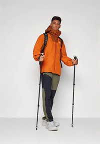 Casaco impermeável laranja com capuz castanho, calças verdes e pretas, e bastões de caminhada. Modelo a usar ténis brancos, de pé sobre um fundo liso.