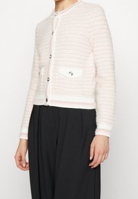 Světle růžový texturovaný cardigan s bílým lemem, který má stříbrné knoflíky a dvě přední kapsy. Měkký pletený materiál, krátký střih.