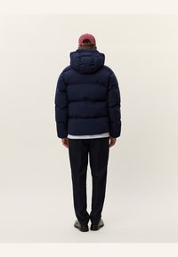 Dunkelblauer Steppjacke mit Kapuze, versehen mit horizontalen Steppnähten, kombiniert mit dunklen Hosen und schwarzen Schuhen, von hinten betrachtet.