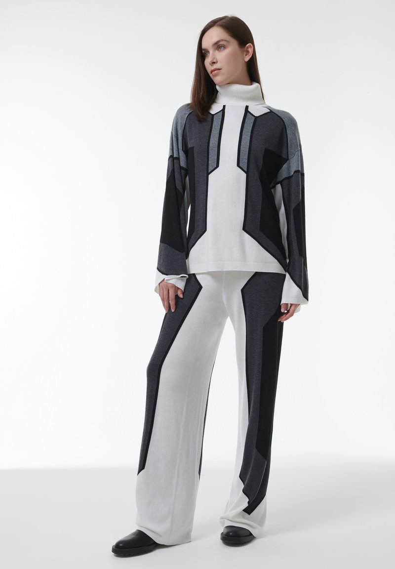 Vrouw in een geometrisch patroon van zwart, grijs en wit turtleneck sweater en bijpassende brede broek met zwarte schoenen, staand op een witte achtergrond.