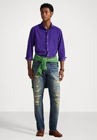 Μπλούζα με κουμπιά μωβ, πράσινο πουλόβερ δέμενο στη μέση, φθαρμένο μπλε τζιν, καφέ παπούτσια. Καθημερινή εμφάνιση με ποικιλία υφών και χρωμάτων.