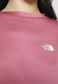 Różowa koszula z krótkim rękawem uszyta z gładkiego materiału, z białym logo "The North Face" na górnej lewej stronie.