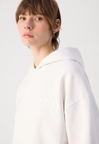 Felpa color beige chiaro con una consistenza morbida e una vestibilità oversize. Presenta un cappuccio con cordino e un logo ricamato tonale sul davanti.