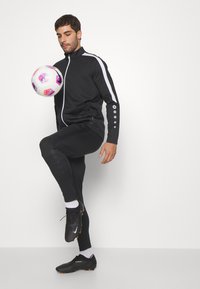Schwarzer sportlicher Trainingsanzug mit weißen Akzenten, bestehend aus einer Reißverschlussjacke und engen Hosen. Schwarze Fußballschuhe, weiße Socken und ein mehrfarbiger Fußball.