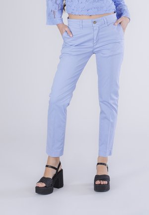 Personne portant un pantalon slim bleu clair et des sandales compensées noires avec des brides à la cheville, les mains dans les poches, debout devant un fond uni.