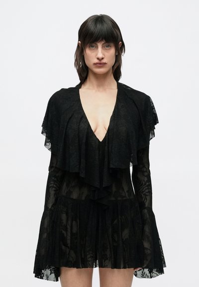 Frau mit dunklen Haaren, die ein durchsichtiges schwarzes Spitzenkleid mit geschichteten Rüschen und tiefem V-Ausschnitt vor einem schlichten weißen Hintergrund trägt.