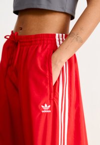 Pantalon de sport rouge avec une taille élastique, des bandes blanches sur les côtés et un petit logo Adidas blanc. Dispose d'une poche latérale et d'un tissu léger.