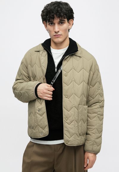 Beige καπιτονέ μπουφάν με διαμαντένιο σχέδιο και ασημί κούμπωμα. Διαθέτει μαύρο στρογγυλό γιακά, το οποίο είναι φορεμένο πάνω από έναν μαύρο πουλόβερ, σ�υνδυασμένο με καφέ παντελόνι.