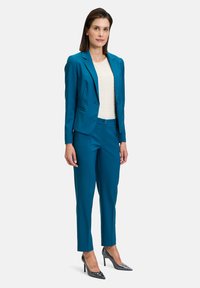 Zöldeskék blézer és hozzáillő nadrág sima anyagból, egy texturált krémszínű toppal párosítva. Zárt orrú fekete magassarkú cipők hangsúlyozzák a szettet.