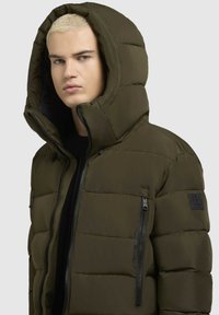 Olivno zeleni napihljeni jopič z velikim kapuco, zadrgo v žepih, pikčasto teksturo in črnimi kovinskimi dodatki.
