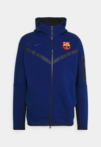 Felpa blu con zip realizzata in un tessuto morbido, caratterizzata da un colletto alto, dettagli neri e un logo del FC Barcelona sul petto.