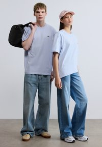 Ανοιχτό μπλε βαμβακερά T-shirt, με χαλαρή εφαρμογή, σε συνδυασμό με φαρδιά μπλε τζιν παντελόνια. Αξεσουάρ περιλαμβάνουν μια μαύρη τσάντα και ένα ροζ καπέλο.