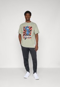 Olivengrønt grafisk T-shirt med en blå tegneseriefigur på en rød og sort eksplosionsbaggrund, parret med mørkegrå sweatpants og hvide sneakers.