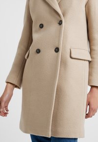 Beige Doppelreiher-Mantel aus strukturierter Fabric, mit schwarzen Knöpfen, einem eingeschnittenen Kragen und seitlichen Taschen, in gerader Silhouette.