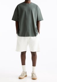 T-shirt ample vert foncé, manches courtes, col rond ; assorti à un short blanc et des baskets marron clair. Style décontracté, coupe ample.