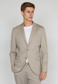 Light beige blazer made of textured fabric, featuring a two-button closure, notched lapels, and front pockets. Worn over a white t-shirt.