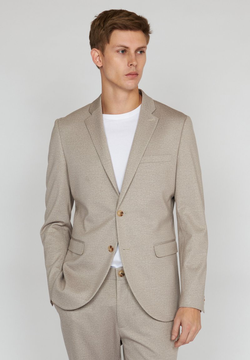 Light beige blazer made of textured fabric, featuring a two-button closure, notched lapels, and front pockets. Worn over a white t-shirt.