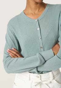 Cardigan a coste di colore azzurro chiaro con scollo rotondo, sei bottoni davanti e maniche lunghe, abbinato a pantaloni bianchi strutturati.