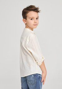 Camicia di lino beige chiaro con maniche arrotolate, abbinata a jeans in denim blu con dettagli sdruciti. Design semplice, vestibilità casual.