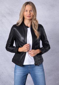 Black leather jacket with a stand collar, front zipper, and side pockets. The material is smooth with a fitted design. Worn over a white top.