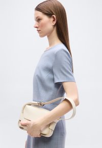 Γυναίκα με ένα κοντομάνικο ανοιχτό μπλε φόρεμα κρατά μια μπεζ τσάντα χειρός από το λουράκι της, μπροστά από ένα απλό φωτεινό φόντο.