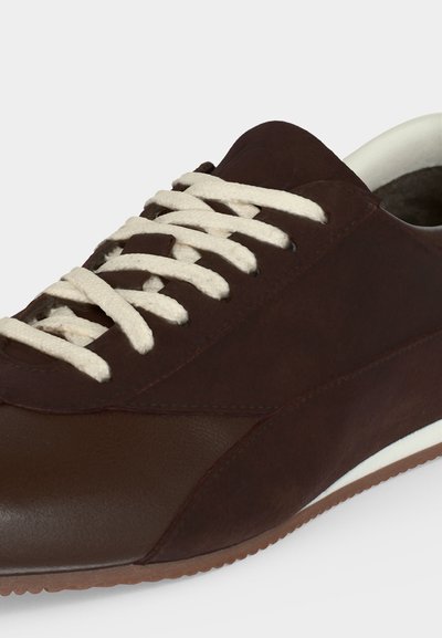 Σκούρο καφέ χαμηλά sneakers με λευκά κορδόνια και λευκή εσωτερική επένδυση σε ανοιχτό φόντο.