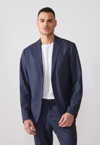Veste de costume bleu marine avec un motif à carreaux texturé, de coupe droite, et des revers crantés. Portée par-dessus une chemise blanche à col rond.