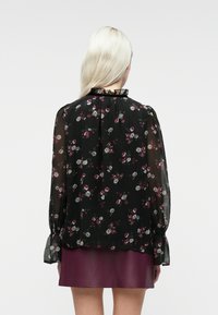 Donna con lunghi capelli biondi, vestita con una blusa nera floreale trasparente e una gonna di pelle viola scuro, di spalle contro uno sfondo semplice.