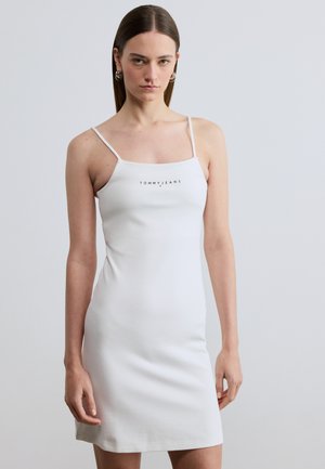 Sieviete valkā pieguļošu balstu bez piedurknēm ar plānām lencītēm un "TOMMY JEANS" logotipu uz krūtīm, stāvot pret vienkāršu fonu.