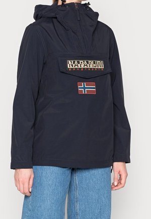 Osoba ubrana w granatową kurtkę Napapijri z przednią kieszenią i naszywką z norweską flagą, w połączeniu z jasnoniebieskimi dżinsami.
