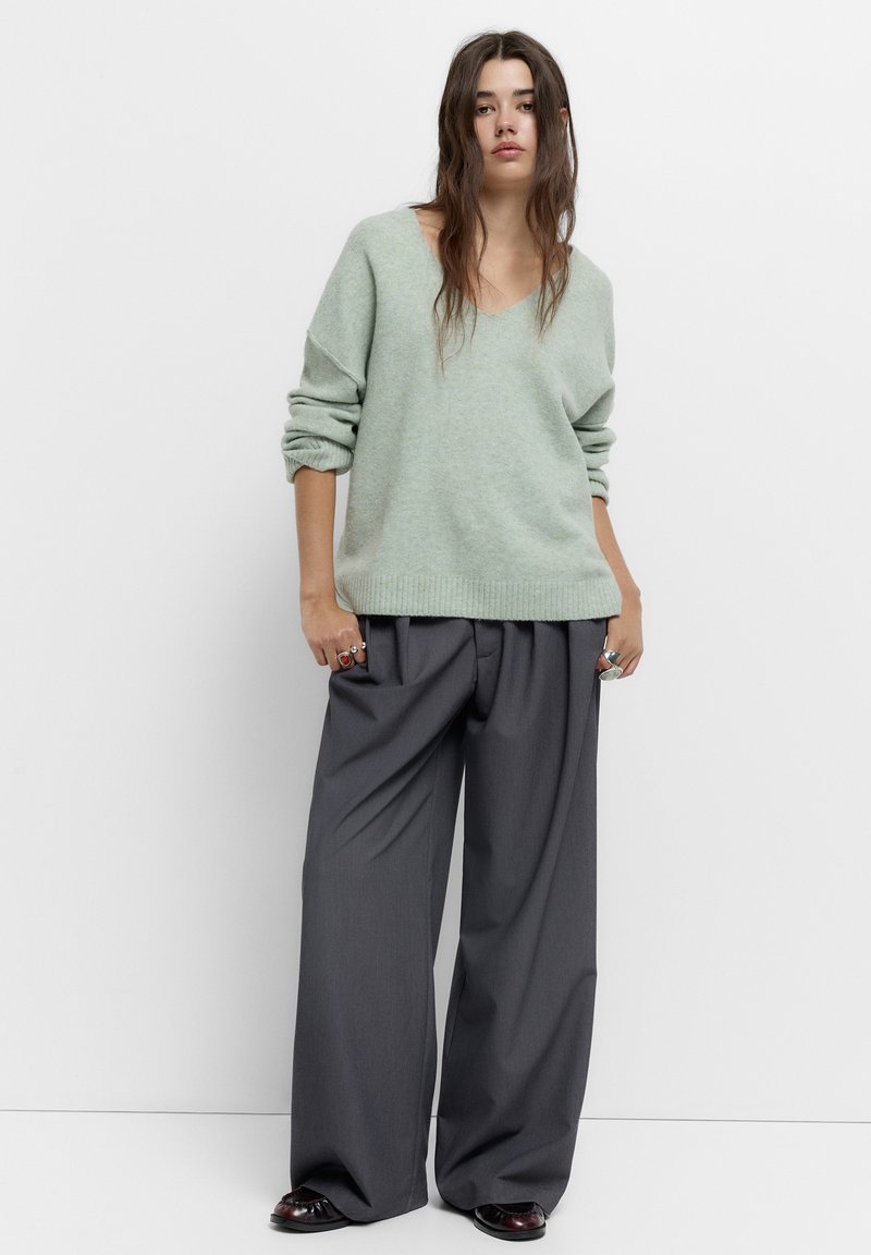 Pulover de lână de culoare verde deschis, cu guler în V, asortat cu pantaloni largi, gri închis, cu pliuri. Textură netedă, cu mansete și tiv ondulate.