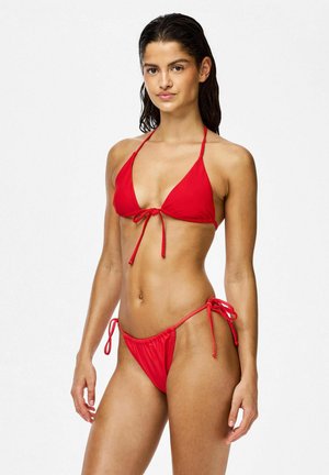 Junge Frau mit nassen dunklen Haaren, die einen roten String-Bikini trägt, steht vor einem schlichten weißen Hintergrund und schaut in die Kamera.