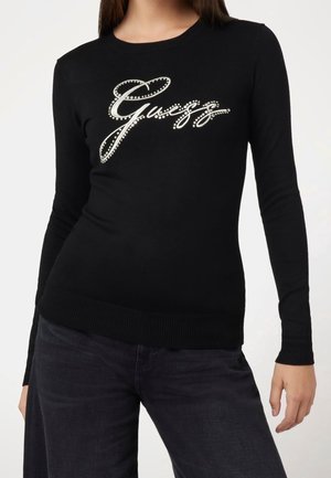 Femme portant un t-shirt noir ajusté à manches longues avec "Guess" écrit en clous blancs sur la poitrine et un pantalon noir.