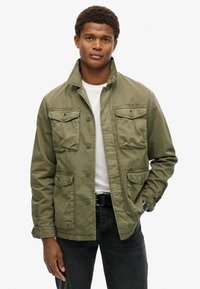 Veste vert olive avec un col, six poches avant, fermeture à boutons et tissu texturé ; portée par-dessus un t-shirt blanc et un jean foncé.