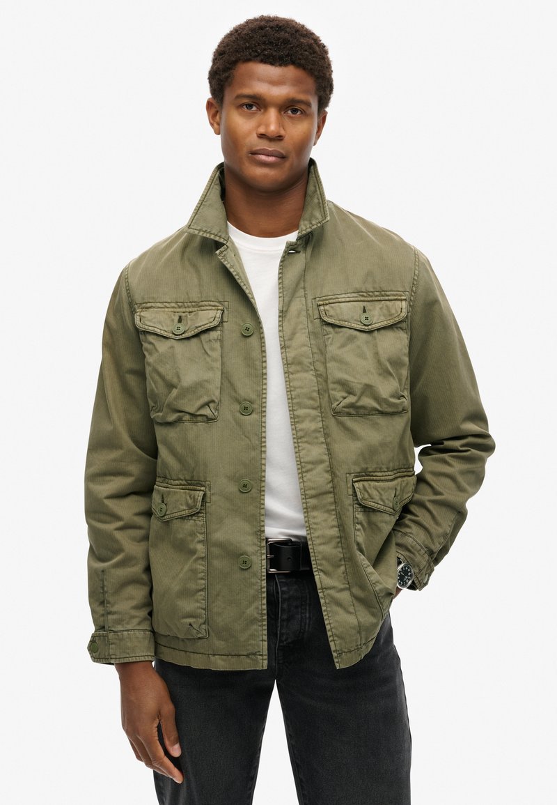 Veste vert olive avec un col, six poches avant, fermeture à boutons et tissu texturé ; portée par-dessus un t-shirt blanc et un jean foncé.