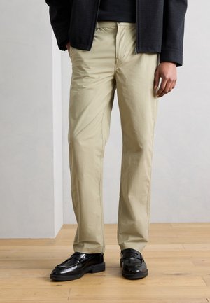 Homme portant un pantalon beige coupe droite, des mocassins noirs et une veste noire, debout sur un sol en bois devant un fond clair uni.