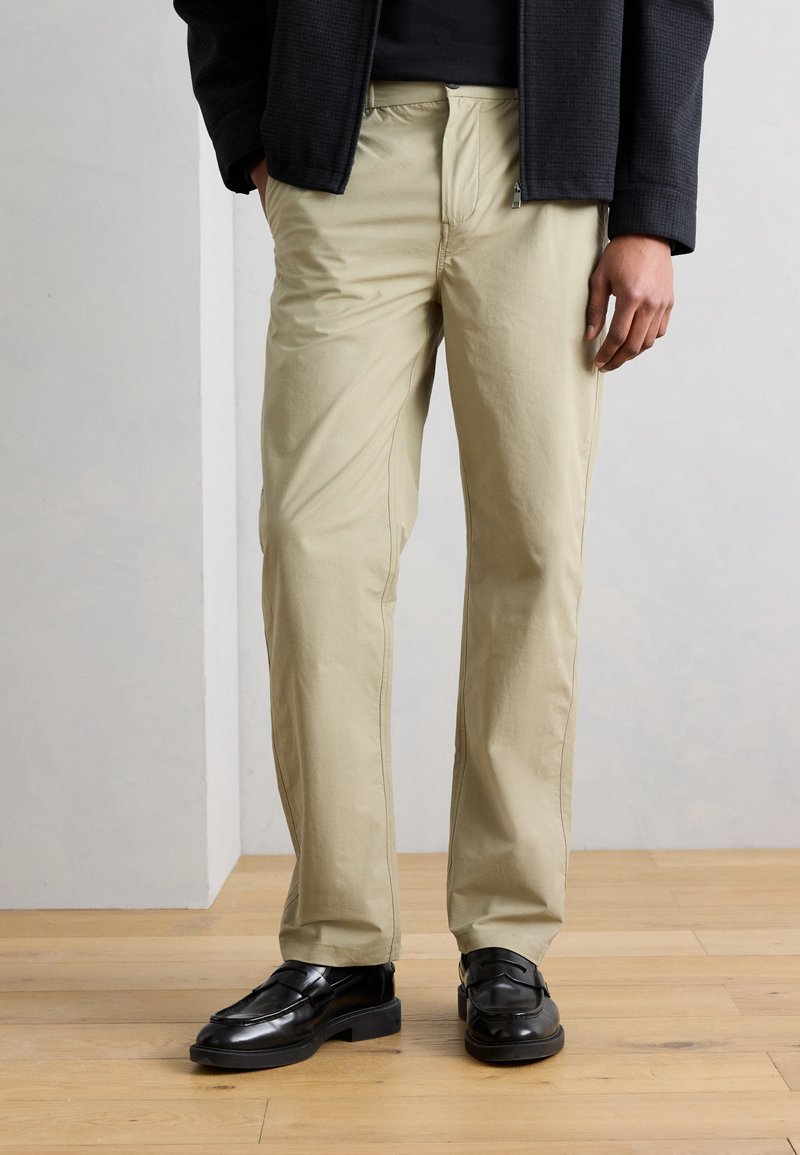 Άνδρας που φοράει μπεζ παντελόνια ευθείας γραμμής, μαύρα μοκασίνια και μαύρο μπουφάν, στέκεται σε ξύλινο πάτωμα μπροστά σε απλό φωτεινό φόντο.