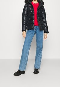 Person klädd i en svart puffjacka med pälsluva, röd logot-skjorta, ljusblå raka jeans och svarta ankelstövlar mot en vit bakgrund.
