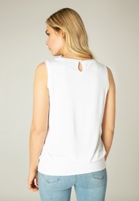 Witte mouwloze top met zachte textuur, sleutelgatdetail op de rug en een ronde halslijn. Gecombineerd met lichtblauwe jeans.